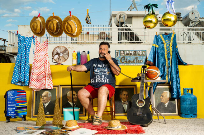 A person sitting on a terrace speaking on an old phone with many things around like paintings, guitar, hats dresses and ore