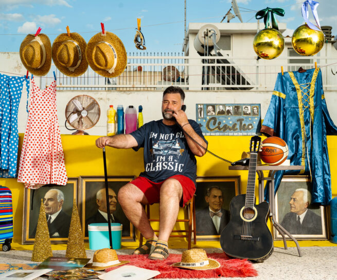 A person sitting on a terrace speaking on an old phone with many things around like paintings, guitar, hats dresses and ore